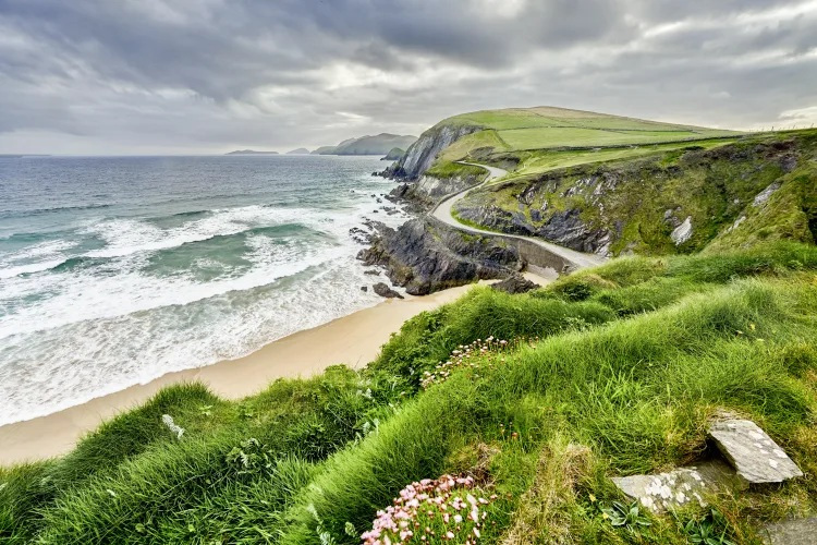 Eine kurvige Küstenstraße schlängelt sich entlang grüner Klippen an einem Sandstrand und dem aufgewühlten Meer unter einem bewölkten Himmel.