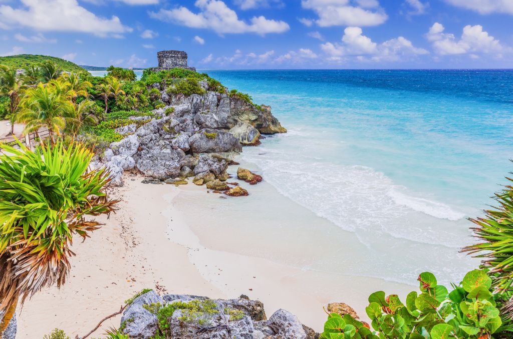 Blick auf die Maya-Ruinen von Tulum auf einer Klippe über einem weißen Sandstrand und türkisblauen Meer. Im Vordergrund Palmen und Felsen. Blick auf die Maya-Ruinen von Tulum auf einer Klippe über einem weißen Sandstrand und türkisblauen Meer. Im Vordergrund Palmen und Felsen.